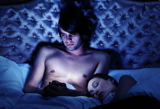 Young couple in bed, woman asleep, man using mobile phone, night